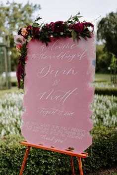 a pink sign with flowers on it sitting in front of a bush and bushes at a wedding