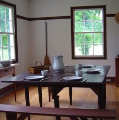 a dining room table with plates and cups on it, near two windows in the background