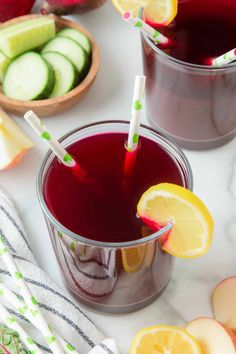 two glasses filled with red liquid next to sliced apples and cucumbers