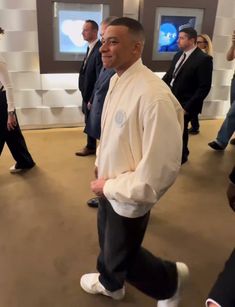 a man in a white jacket and black pants walking through a room with other people