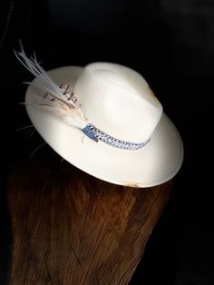 Felt hat with accent bands, matching feather bundle with complimenting details around the hat. This cream colored hat has a mainly blue accent hat band wrapped with a silver, thinner, band to give some shine to the overall appearance.  The feather bundle is wrapped with denim blue fabric to match the hat band. This same denim is also sewn onto the back of the hat for added accents. Brown wax thread was used to sew the denim patch.  Various areas around the hat and brim are burned for an added rustic/ grunge look. A special detail: you will find two small chains hanging from the front left of the brim to match the silver hat band. Adjustable Fedora With Feather Trim For Kentucky Derby, Custom Wide Brim Hat With Feathers, Feathered Brimmed Hat Bands For Summer, Bohemian Fedora Hat Band With Feather Trim, Feathered Hat Bands With Curved Brim For The Beach, Feathered Hat Bands For Beach With Curved Brim, Brimmed Hat Bands With Feathers For Summer, Festival Fedora Hat Band With Feather Trim, Artisan Hats With Feathers And Adjustable Fit