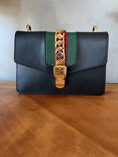 This gently used Authentic Gucci Sylvie shoulder bag has been expertly crafted from black leather with a gold tone chain web decorated flap and buckle lock accented with Gucci's signature green red stripe. It has a pristine tan suede interior and green and red striped fabric strap. The purse was barely used and the only sign of wear is on the turn lock closure, buckle, front decorative chain, and gold toned accents on strap. It comes without a dust bag or original box. Designer Satchel With Chain Strap, Designer Chain Strap Satchel Shoulder Bag, Designer Flap Shoulder Bag With Chain Strap, Designer Shoulder Flap Bag With Chain Strap, Designer Green Flap Bag With Gold-tone Hardware, Gucci Sylvie, Fabric Strap, Striped Fabric, Green And Red