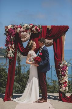 a bride and groom standing under an arch decorated with red, white and pink flowers