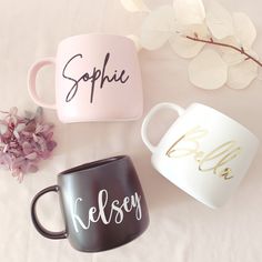 three coffee mugs sitting on top of a bed next to purple flowers and leaves