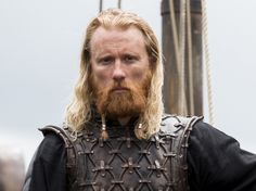 a man with long blonde hair and beard wearing armor is standing in front of a building