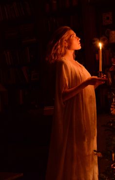 a woman holding a lit candle in her hand while standing next to a book shelf