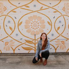 a woman kneeling down in front of a wall with flowers painted on it and a flowered design behind her