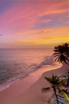 the sun is setting over the ocean with palm trees and lounge chairs on the beach