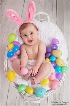 a baby wearing bunny ears sitting in a basket filled with easter eggs and candies