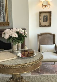 a table with flowers and books on it in front of a mirror, chair and fireplace