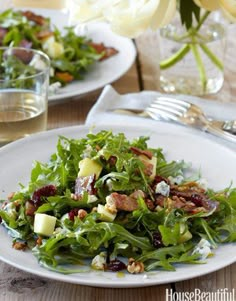 two white plates with salad on top of them next to wine glasses and silverware