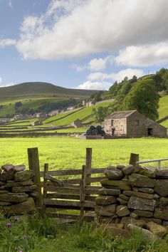 a stone wall and gate in front of a green field with hills behind it,