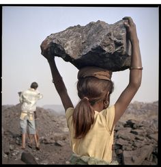 a woman carrying a rock on her head while another man walks by in the background