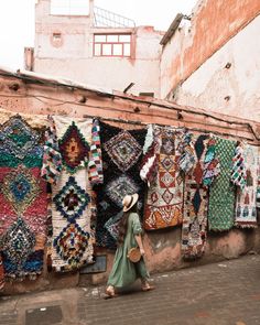 a woman walking down a street next to a wall covered in rugs and blankets