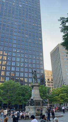 many people are standing around in front of tall buildings and one person is sitting on a bench