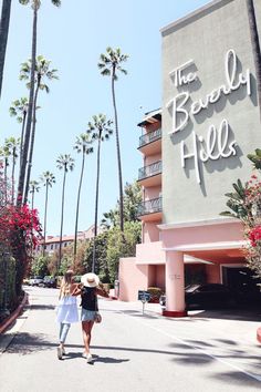 two women walking down the street in front of a hotel with palm trees on both sides