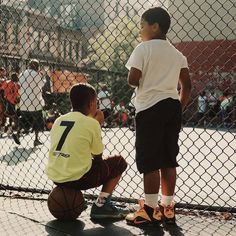 two young boys are playing basketball on a court while others watch from behind a chain link fence