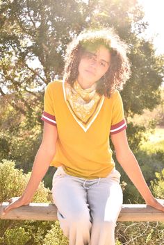 Woman wearing block print Tate bandana around the neck against a yellow tee shirt. Woman sitting on wooden fence with trees in the background. Casual Patterned Scarves For Summer, Summer Cotton Patterned Bandana, Casual Patterned Bandana For Festivals, Patterned Casual Bandana For Festival, Trendy Cotton Summer Scarves, Trendy Summer Cotton Scarves, Multicolor Cotton Bandana, Casual Cotton Patterned Bandana, Trendy Cotton Scarves For Summer