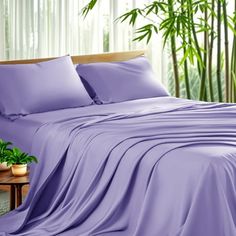 a bed covered in purple sheets and pillows next to a plant on a wooden table