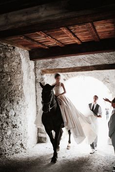 a woman riding on the back of a black horse in a stone walled area with people standing around