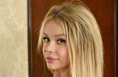 a woman with long blonde hair and blue eyes looking at the camera while standing in front of a door