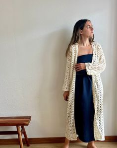 a woman standing in front of a white wall wearing a long cardigan and blue dress