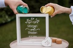 two people are holding cans and pouring them into a photo frame on a small table