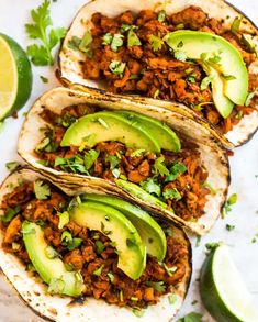 three tacos filled with meat, avocado and cilantro on a white surface