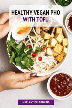 two hands holding a bowl of food with chopsticks and sauce on the side