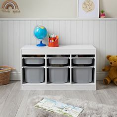 a white shelf with several bins and a teddy bear on the floor next to it