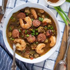 a bowl of soup with shrimp, sausage and green onions on a blue towel next to two spoons