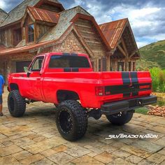 two men standing next to a red truck in front of a large wooden house on a cloudy day