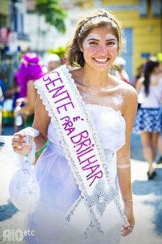 a woman in a white dress holding a pink sash and smiling at the camera with other people behind her