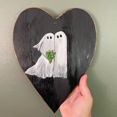 a hand holding up a painted wooden heart shaped like two ghostes with flowers in their hands