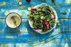 a bowl of salad next to a cup of dressing on a blue wooden table top