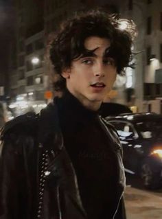 a young man with curly hair wearing a black leather jacket in the city at night