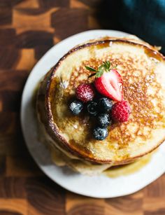 pancakes with berries and powdered sugar sit on a white plate atop a wooden table