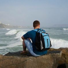 a young man sitting on top of a rock near the ocean looking out to sea