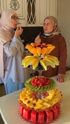 two women standing next to each other in front of a cake with fruit on it