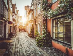 an old cobblestone street with tables and chairs in the sun shining through windows