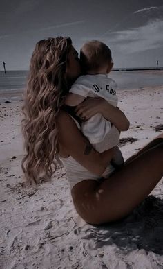 a woman sitting on top of a sandy beach next to the ocean holding a baby