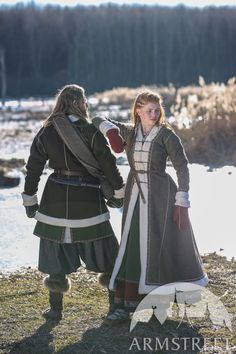 two people dressed in medieval clothing standing next to each other on the grass near water