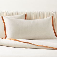 a bed with white sheets and orange trim on the pillowcase, in front of a beige headboard