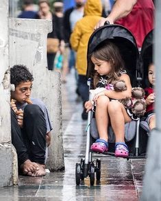 two young children sitting in strollers on the sidewalk while people walk down the street