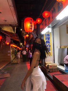 a woman in a white dress walking down a street with lanterns hanging above her head