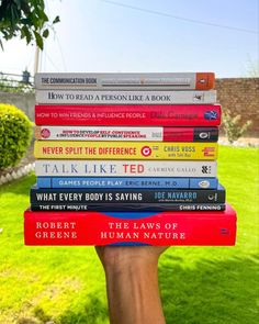 a hand holding a stack of books in front of a green lawn with trees and bushes