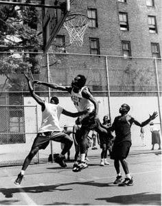 black and white photograph of young men playing basketball in the street with apartment buildings behind them