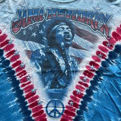 Step into the music scene with this Vintage Jimi Hendrix American Flag Blues Guitarist Tie Dye Tee. Officially licensed and medium-sized, this shirt is a genuine tribute to the legendary guitarist. Its tie-dye pattern with the American flag motif channels the hippie vibes of the era. Ideal for music enthusiasts, collectors, and those who appreciate vintage fashion, this tee is more than apparel; it's a piece of concert memorabilia representing a crucial moment in music history. Whether you're a Pop Culture Tops With Band Logo For Music Festivals, Pop Culture Crew Neck Top For Music Festivals, Rock And Roll Crew Neck Tops For Music Festival, Festival Cotton Tops With Band Logo, Rock And Roll Screen Print Tops For Music Festivals, Rock And Roll Cotton Top For Music Festival, Band Logo Cotton Tops For Festival, Blue T-shirt For Summer Concerts, Cotton Band Logo Tops For Festival