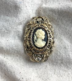 "You will receive this vintage cameo brooch from Gerry's. It measures 1 3/4\" x 1 1/8\" and is cast in an antiqued gold tone base metal. The cameo backing is black and the silhouette is a cream color. This brooch is in very good vintage condition with minimal signs of wear and a properly functioning safety catch. For finished vintage assemblage and vintage inspired necklaces, bracelets, and earrings, please visit us at our other Etsy store, William Dalton Design: www.williamdaltondesign.etsy.com Vintage Gold Brass Brooches, Antique Gold Cameo Brooches, Gold Cameo Brooch Collectible, Gold Antique Brooches For Vintage Collection, Antique Gold Cameo Brooch, Antique Gold Brooches For Vintage Collection, Antique Gold Brooches From Vintage Collection, Vintage Filigree Brooches, Victorian Gold Brass Brooches