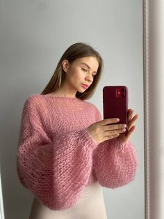 a woman wearing a pink sweater looking at her cell phone while standing in front of a mirror